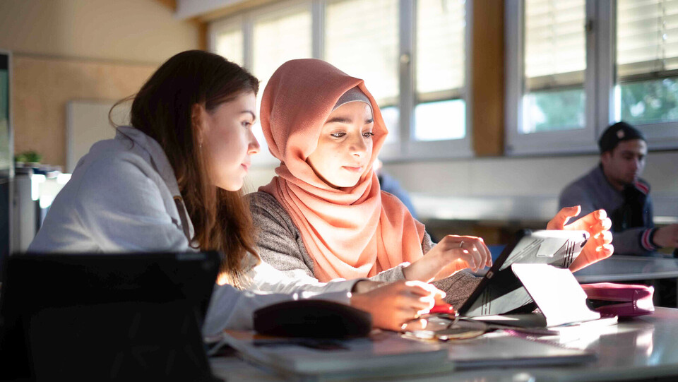 Zwei junge Frauen sitzen in einem Seminarraum und interagieren mit einem Tablet