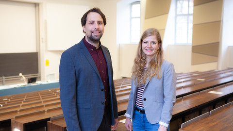 Zwei Personen in schicker Kleidung stehen in einem leeren Hörsaal. Die Personen heißen Tobias Ebert und Jana Berkessel.