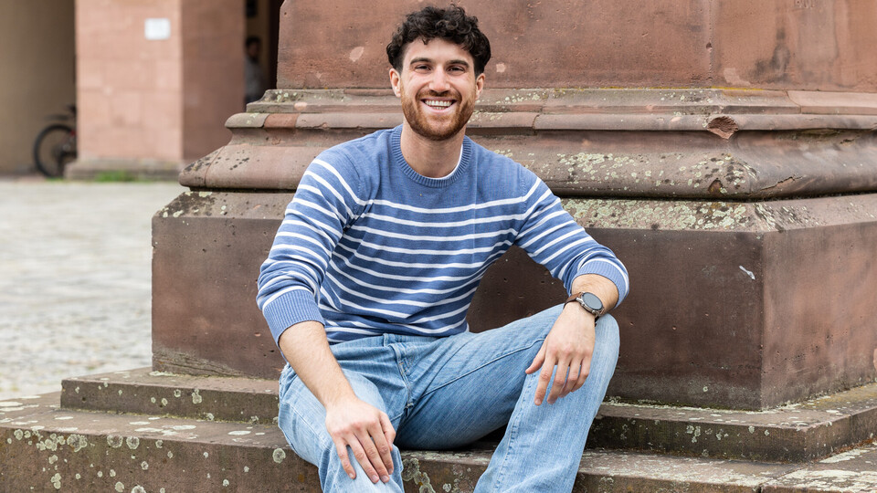 Marco sitzt auf den Stufen einer Statue auf dem Ehrenhof. Er trägt eine blaue Jeans und einen blau-weiß-gestreiften Pullover.