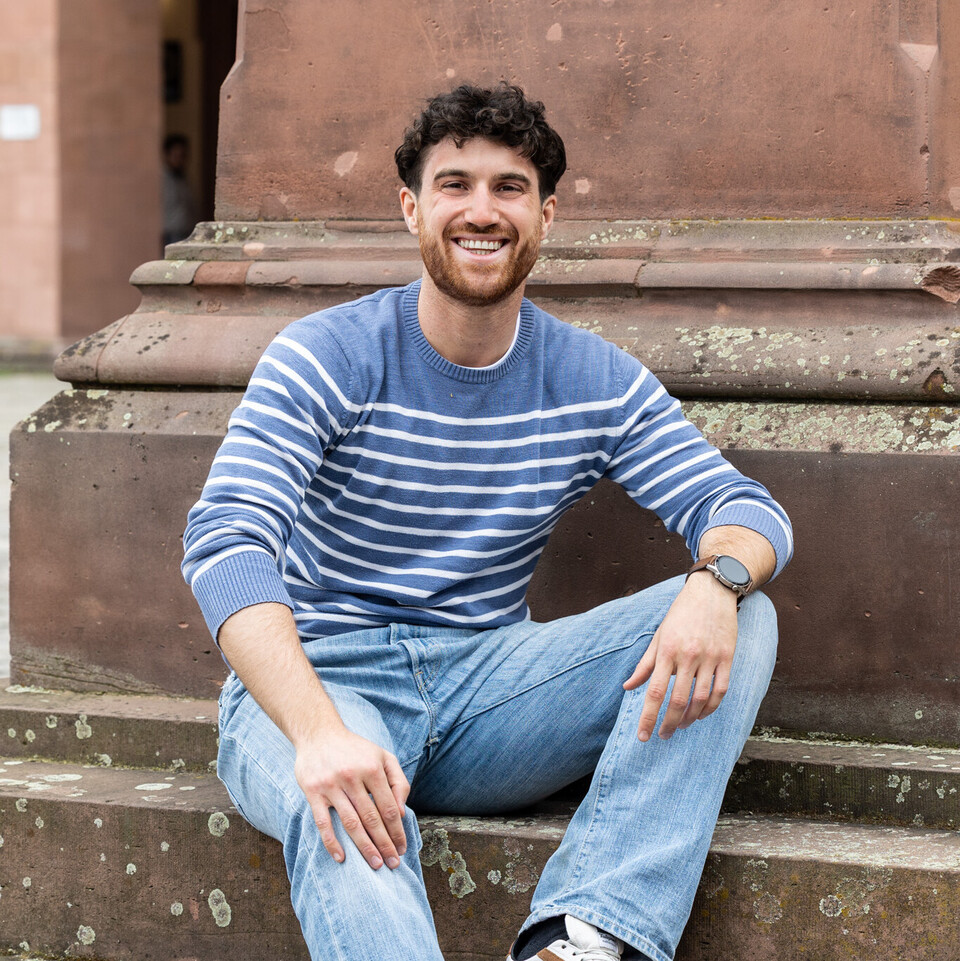 Marco sitzt auf den Stufen einer Statue auf dem Ehrenhof. Er trägt eine blaue Jeans und einen blau-weiß-gestreiften Pullover.