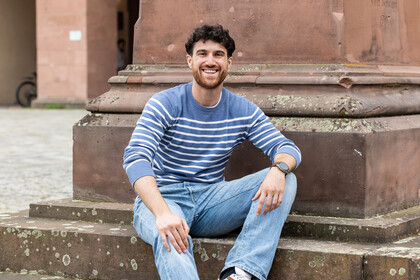 Marco sitzt auf den Stufen einer Statue auf dem Ehrenhof. Er trägt eine blaue Jeans und einen blau-weiß-gestreiften Pullover.