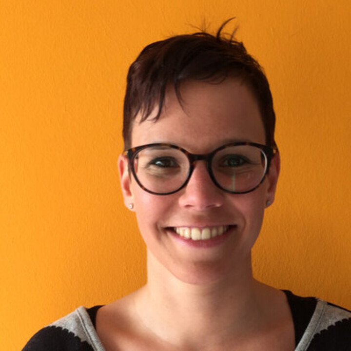 Portrait von Christiane Grill. Sie trägt ein schwarz-grauen Pullover und steht vor einer orange-farbigen Wand.