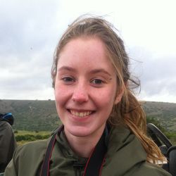 Jessica Horn hat vom Wind zerzauste Haare und lächelt in die Kamera. Sie trägt eine olivgrüne Jacke. Im Hintergrund die grüne Natur Südafrikas.