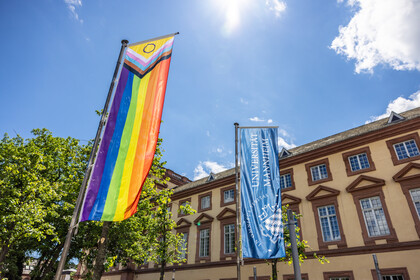 Vor dem Ostflügel des Mannheimer Schlosses wurde die Progress Pride Flag gehisst. Sie wird auch erweiterte Regenbogenflagge genannt. Daneben weht die dunkelblaue Fahne der Universität Mannheim.