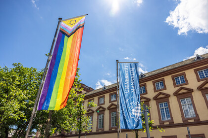 Vor dem Ostflügel des Mannheimer Schlosses wurde die Progress Pride Flag gehisst. Sie wird auch erweiterte Regenbogenflagge genannt. Daneben weht die dunkelblaue Fahne der Universität Mannheim.