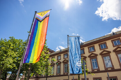 Vor dem Ostflügel des Mannheimer Schlosses wurde die Progress Pride Flag gehisst. Sie wird auch erweiterte Regenbogenflagge genannt. Daneben weht die dunkelblaue Fahne der Universität Mannheim.