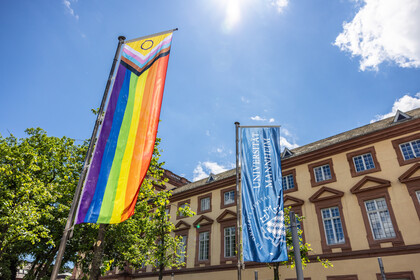 Vor dem Ostflügel des Mannheimer Schlosses wurde die Progress Pride Flag gehisst. Sie wird auch erweiterte Regenbogenflagge genannt. Daneben weht die dunkelblaue Fahne der Universität Mannheim.