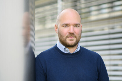 Porträt von Prof. Traunmüller. Er trägt einen dunklen Pullover und lehnt an einer Säule.