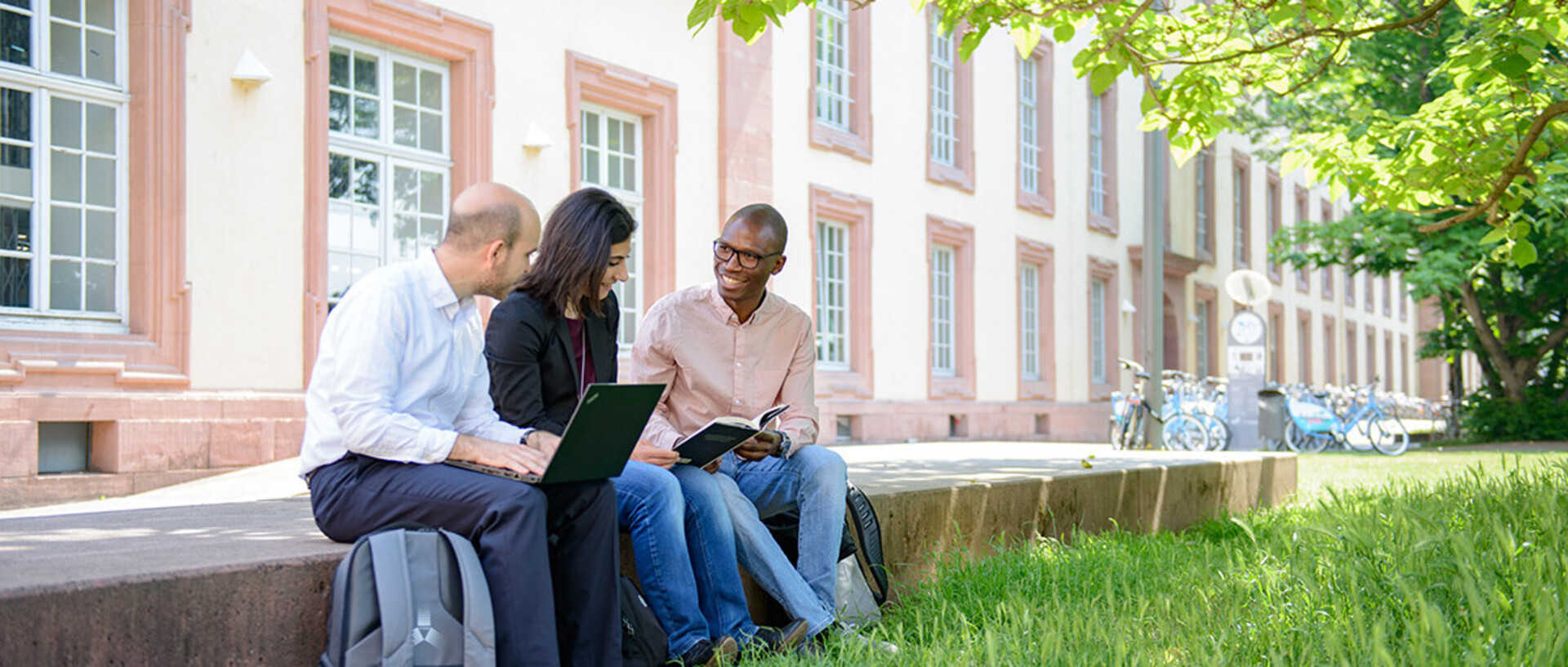 Three people are sitting on a bench in front of the palace of the University of Mannheim. Suitcases and bags are placed beside them. In the foreground, there is a lawn with flowering plants. The scene takes place on campus on a sunny day.