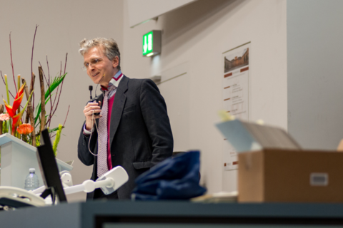 Prof. Dr. Justus Fetscher spricht in eiin Mikrophon. Neben ihm ein Blumengesteck.
