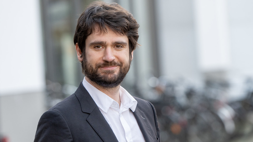 Marius Sältzer. He is wearing a black suit and is standing outside a tall building.