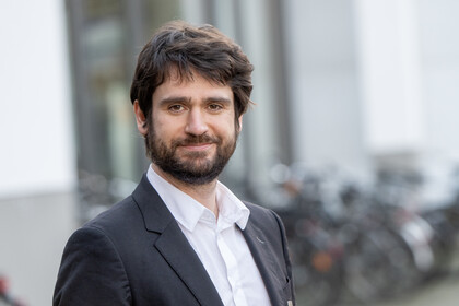 Marius Sältzer. He is wearing a black suit and is standing outside a tall building.