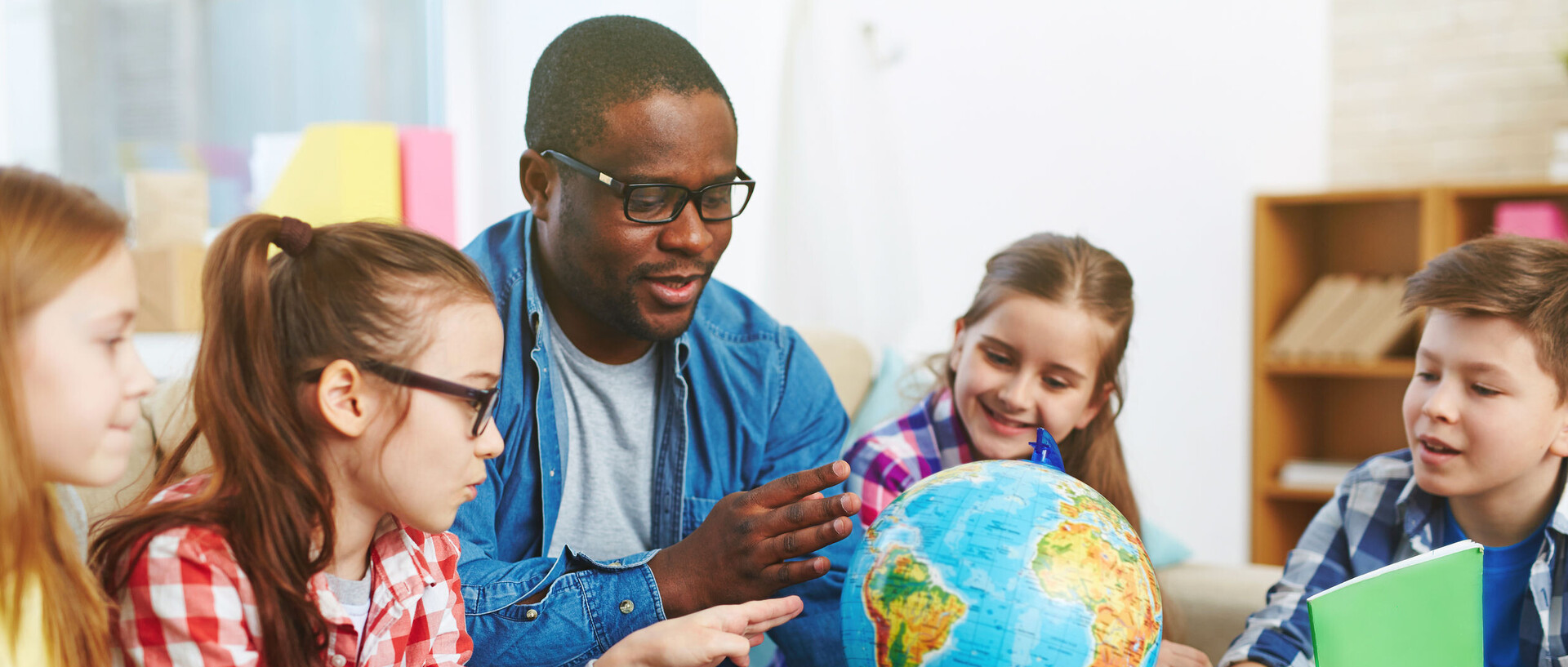 Ein Mann zeigt Hält einen Globus in seiner Hand und zeigt auf ein Land. Vier Kinder schauen ihm interessiert dabei zu.