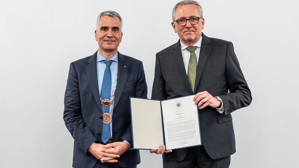 Peter Kurz trägt einen dunkelgrauen Anzug mit grüner Krawatte, Brille und kurze Haare. Er erhält die Ehrensenatorwürde von Prof. Dr. Thomas Fetzer, der einen dunkelblauen Anzug mit blauer Krawatte trägt. Beide stehen nebeneinander und lächeln in die Kamera.