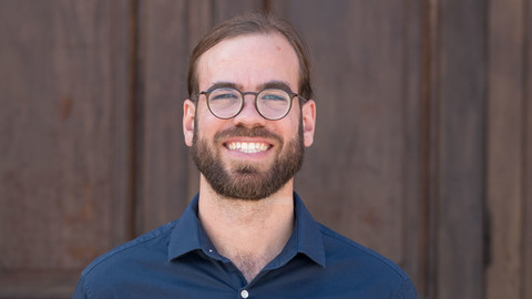 Daniel Overbeck. He is wearing a blue shirt and is standing in front of a brown door.