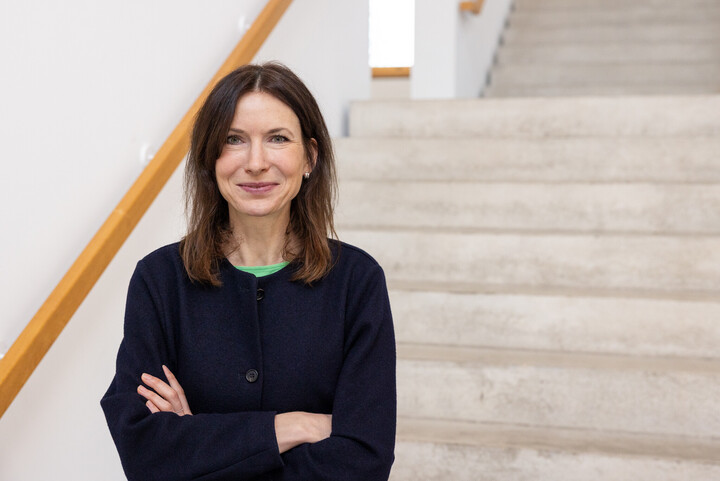 Porträtbild von Sarah Geber. Sie steht vor einer Treppe und verschränkt die Arme.