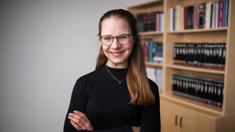 Laura Montenbruck with brown glasses, smiling with arms crossed, wearing a black turtleneck, with a blurred bookshelf in the background.