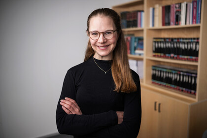 Laura Montenbruck with brown glasses, smiling with arms crossed, wearing a black turtleneck, with a blurred bookshelf in the background.
