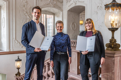 Drei Personen lächeln in die Kamera. Sie stehen nebeneinander. Zwei zeigen eine Urkunde.