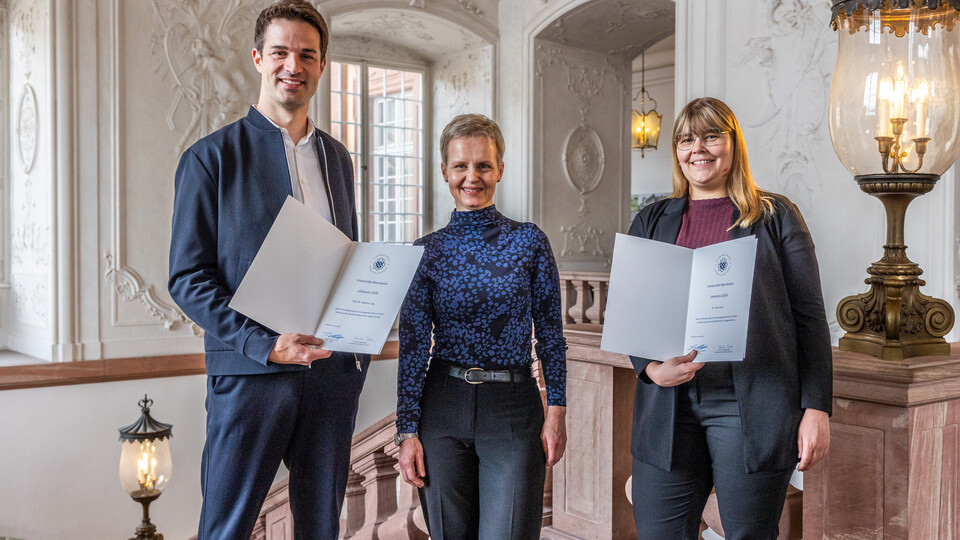 Drei Personen lächeln in die Kamera. Sie stehen nebeneinander. Zwei zeigen eine Urkunde.