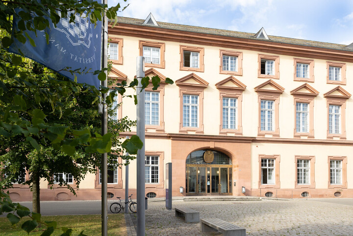 Blaue Flagge mit dem Logo der Universität Mannheim vor dem Haupteingang der Universität Mannheim.