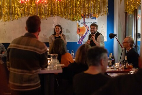 Viele Personen sitzen in einer Bar und schauen zu einer Person, die mit einer Mikrofon in der Hand spricht.