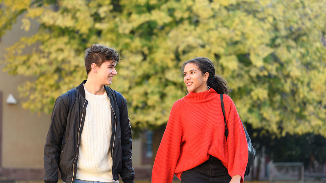 Eine Studentin und ein Student spazieren nebeneinander her und unterhalten sich. Die Studentin trägt einen roten Pullover. Der Student trägt einen weißen Strickpullover und darüber eine schwarze Jacke.