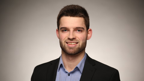 Jakob Schmidhäuser. He is wearing a black suit and is standing in front of a brownish wall.