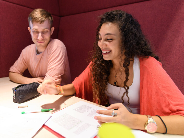 Zwei Studierende sitzen lachend an einem Tisch. Vor ihnen ausgedruckte Texte, Stifte und ein Kaffeebecher.