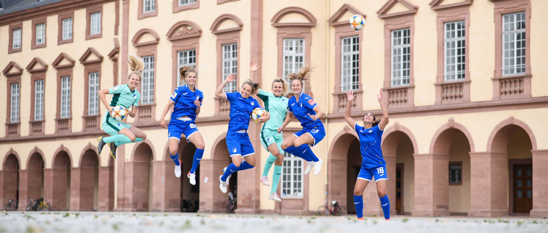 Sechs Fußballerinnen springen, in blauen Trikots gekleidet, vor dem Mannheimer Schloss in die Luft.