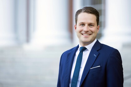 Gunther Glenk trägt einen blauen Anzug mit einer blauen Krawatte und lächelt.