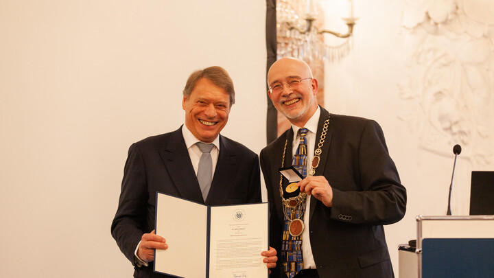 Dr. John Feldmann trägt einen Anzug und hält die offene Ehrenwürdigung in der Hand. Neben ihm steht Prof. Dr. Puhl ebenfalls im Anzug und einer goldenen Medaille in der Hand. Im Hintergrund die Marmorwand des Rittersaals der Uni Mannheim.