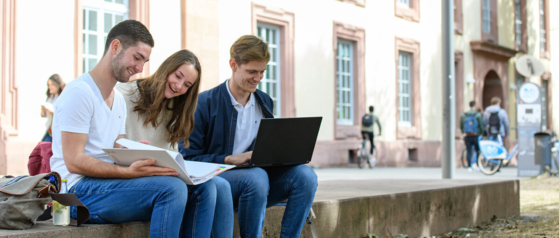 Studierende sitzen vor dem Schloss auf einer Mauer. Sie haben Laptops oder Bücher, in die sie schauen.