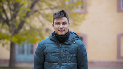 Eine Person trägt eine dunkle türkisfarbene Jacke sowie einen schwarzen Schal und steht vor dem Schloss. Die Person heißt Oleksandr Kova.