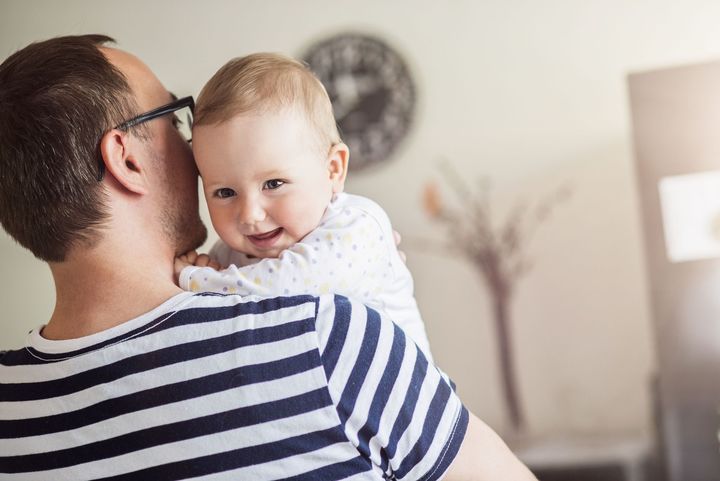 Mann mit blau weiß gestreiften T-Shirt hat Baby auf dem Arm. Baby lächelt in die Kamera und hat einen weißen Body mit gelb-blauen Punkten an.