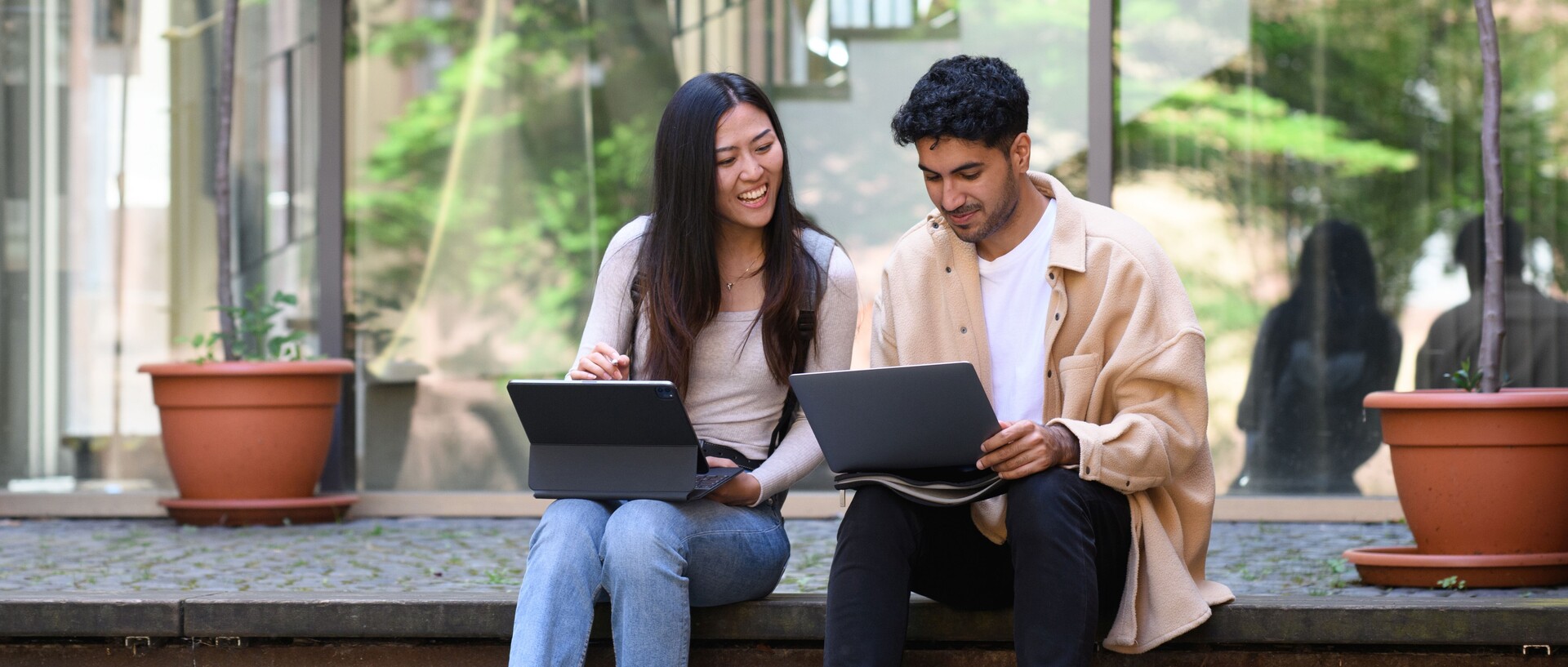 Eine Studentin und ein Student sitzen lachend auf einer Treppe und arbeiten an ihren Laptops. Link: Informationen zum Master-Infotag