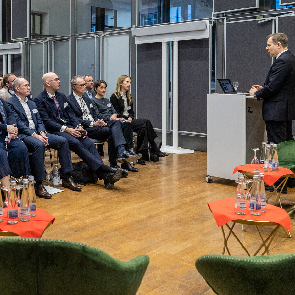 Dutzende Personen sitzen in einem Raum und hören einer Person zu, die am Podium steht.