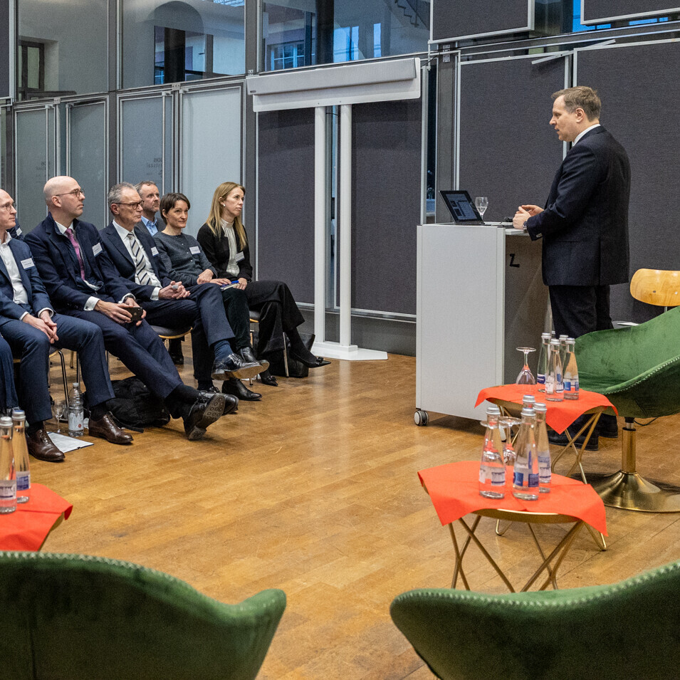 Dutzende Personen sitzen in einem Raum und hören einer Person zu, die am Podium steht.