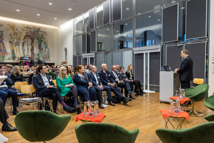 Dutzende Personen sitzen in einem Raum und hören einer Person zu, die am Podium steht.