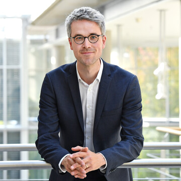 Portait of Prof. Dr. Manuel Trenz. He is wearing a white shirt with a dark blue jacket over it and is standing leaning against a banister.