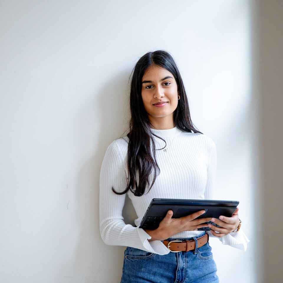 Eine junge Frau steht vor einer Wand. Sie hält ein Tablet in der Hand und lächelt in die Kamera.
