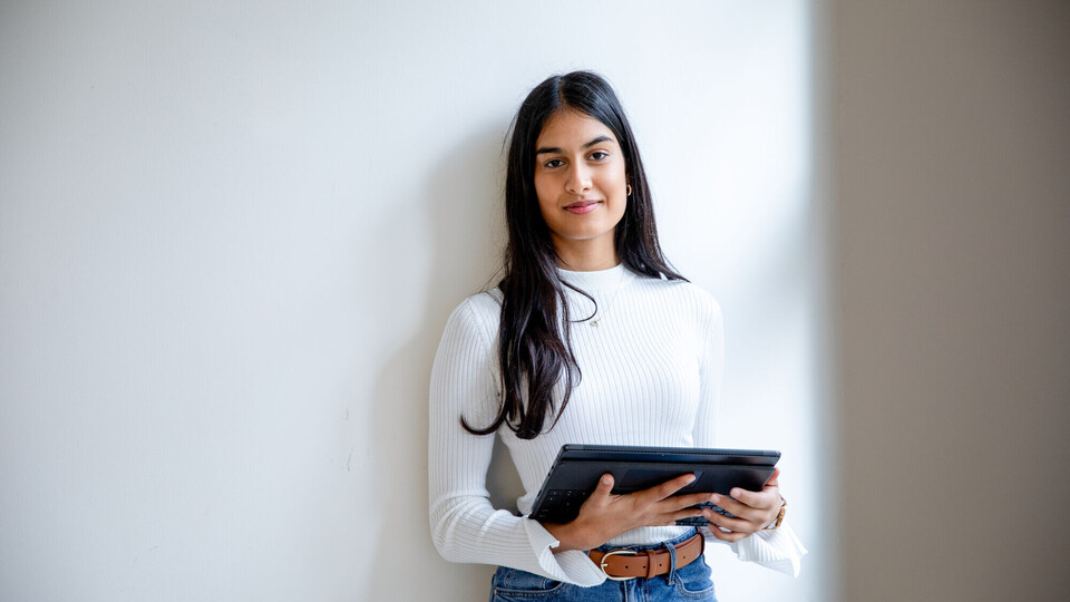 Eine junge Frau steht vor einer Wand. Sie hält ein Tablet in der Hand und lächelt in die Kamera.
