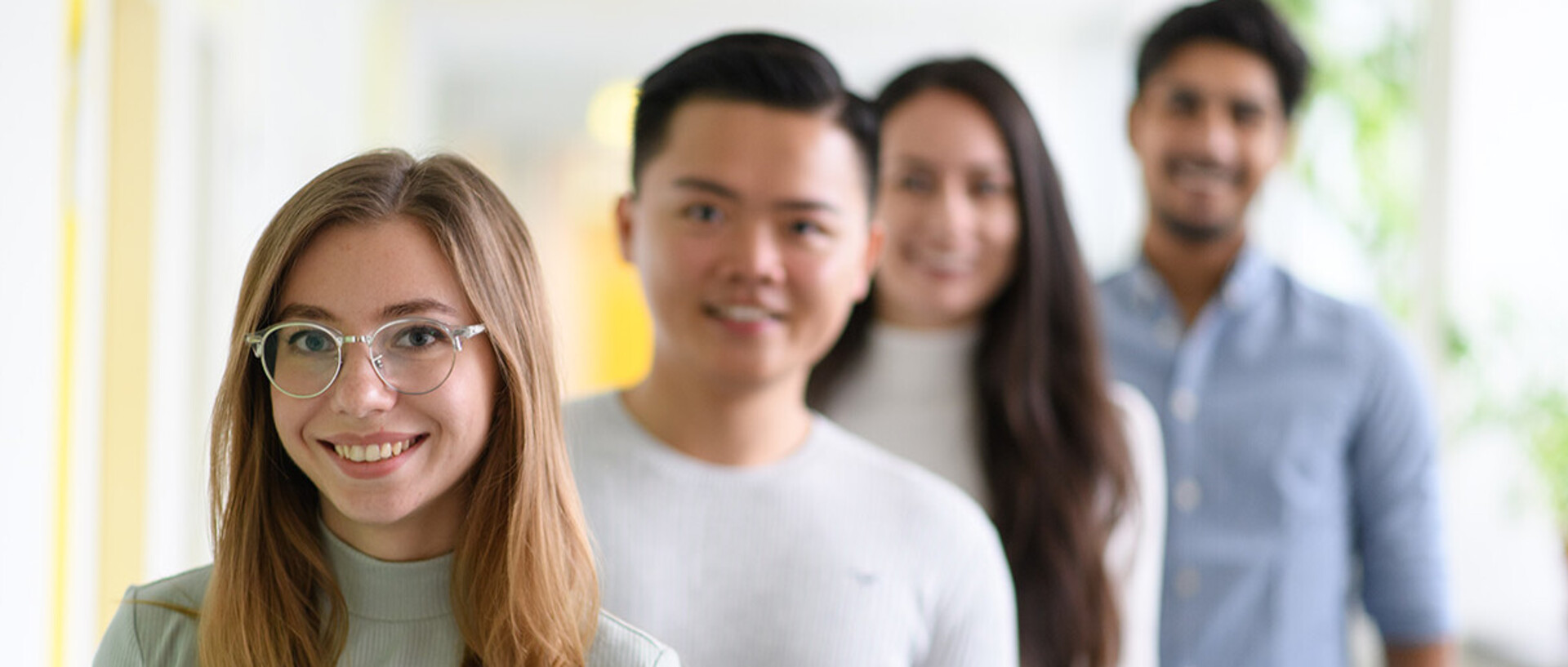Vier Studierende stehen in einer Reihe versetzt hinterheinander und lachen in die Kamera. Nur die Studentin an vorderster Stelle ist scharf gestellt.