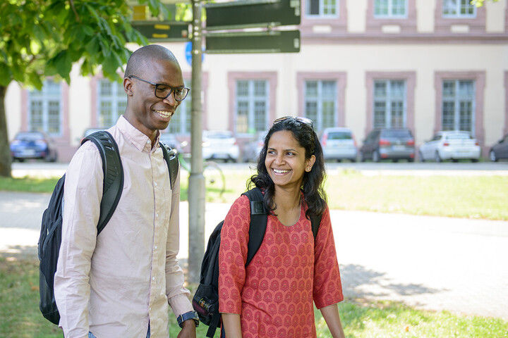 Zwei Personen laufen lachend über das Campus-Gelände. Sie tragen beide schwarze Rucksäcke.