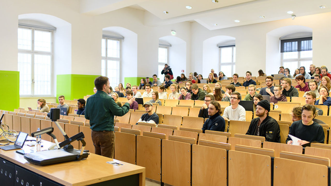 Studierende sitzen in einem Hörsaal. Ein Dozent hält einen Vortrag und gestikuliert mit seiner Hand.