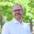 Eine Person trägt ein weißes Hemd, eine blaue Jeans sowie einen braunen Gürtel und steht vor grünen Bäumen. Die Person hat ihre Daumen in die Hosentaschen gesteckt und heißt Jochen Gebauer.