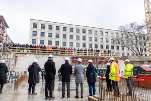 Aufnahme der Grundsteinlegung des A5-Gebäudes der Uni Mannheim. Arbeitende stehen mit weißen Helmen unten, während oben ein Publikum zuschaut.