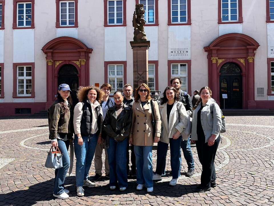 Eine Gruppe von 9 Personen steht vor einem Brunnen auf einem gepflasterten Platz vor einem historischen Gebäude.