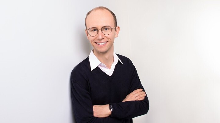Tobias Kalmbach. He is wearing a black sweater and is standing in front of a white wall.