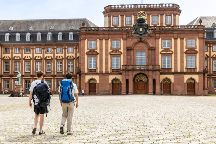 Zwei Studierende mit Reiserucksäcken auf dem Ehrenhof vor dem Schloss.